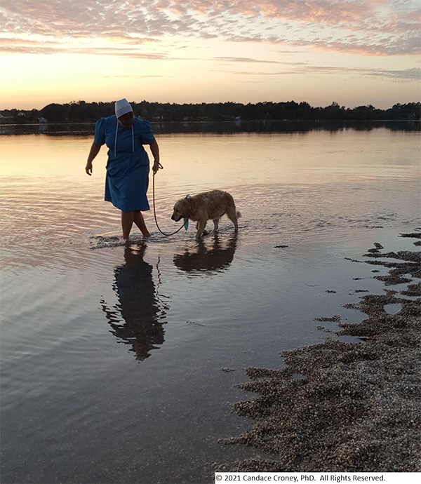 An Amish woman walks an older golden retriever on a leash at sunset in the water along the edge of a lake. 
